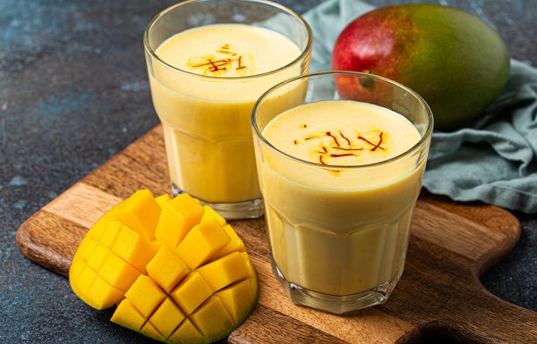 Healthy Indian Ayurveda drink mango lassi in two glasses on rustic concrete table with fresh ripe cut mango, yellow blended beverage made of mango fruit, yoghurt or milk curd and spices